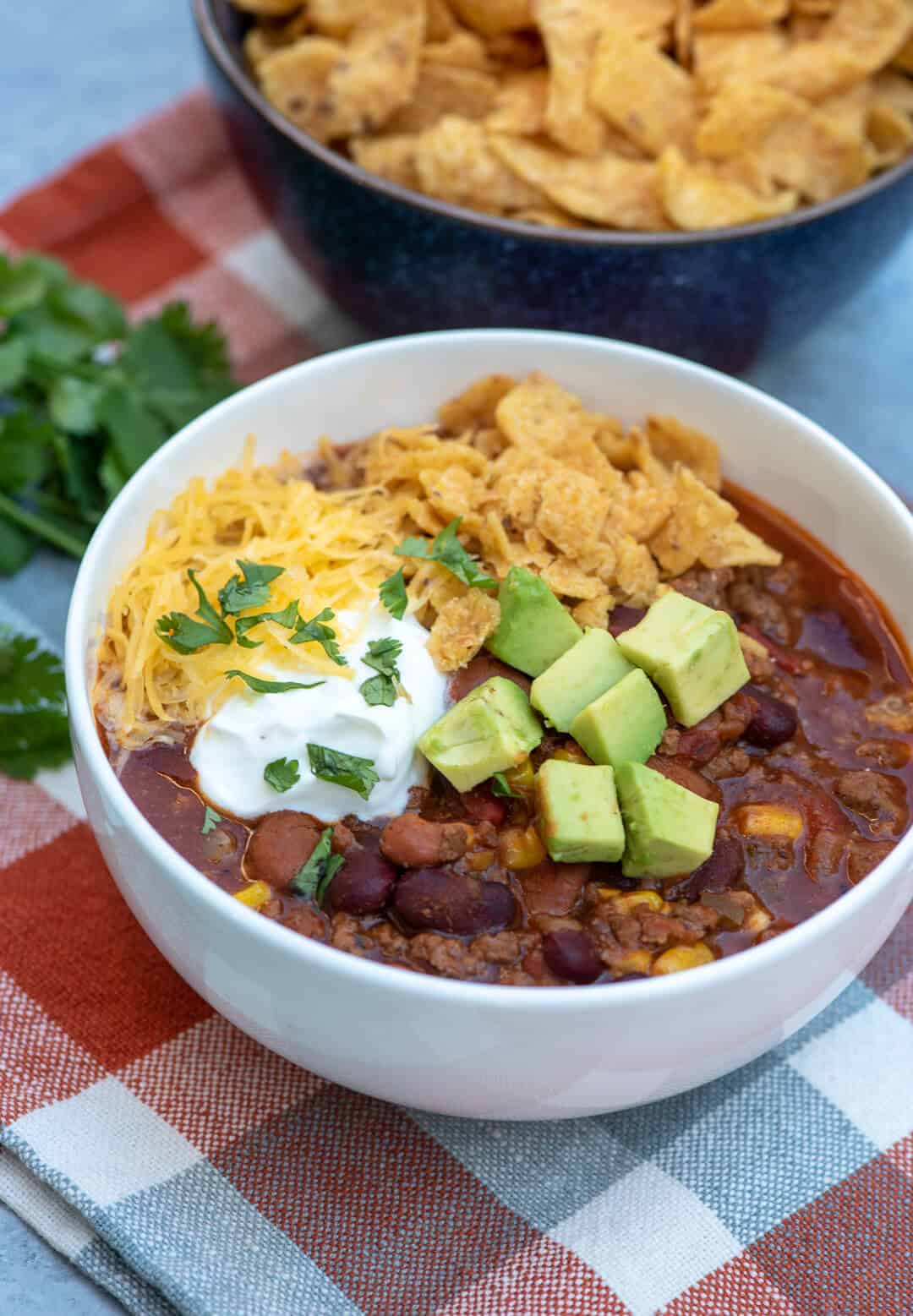 A serving of Beef and Bean Taco Chili in a white bowl garnished with corn chips, sour cream, avocado and cheese on a checked cloth.