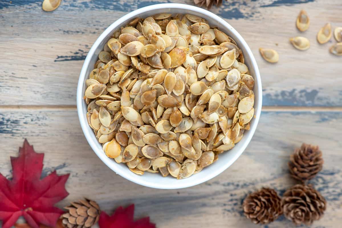 A top down shot of a bowl of roasted acorn squash seeds on a brown ball with fall leaves and pine cones.