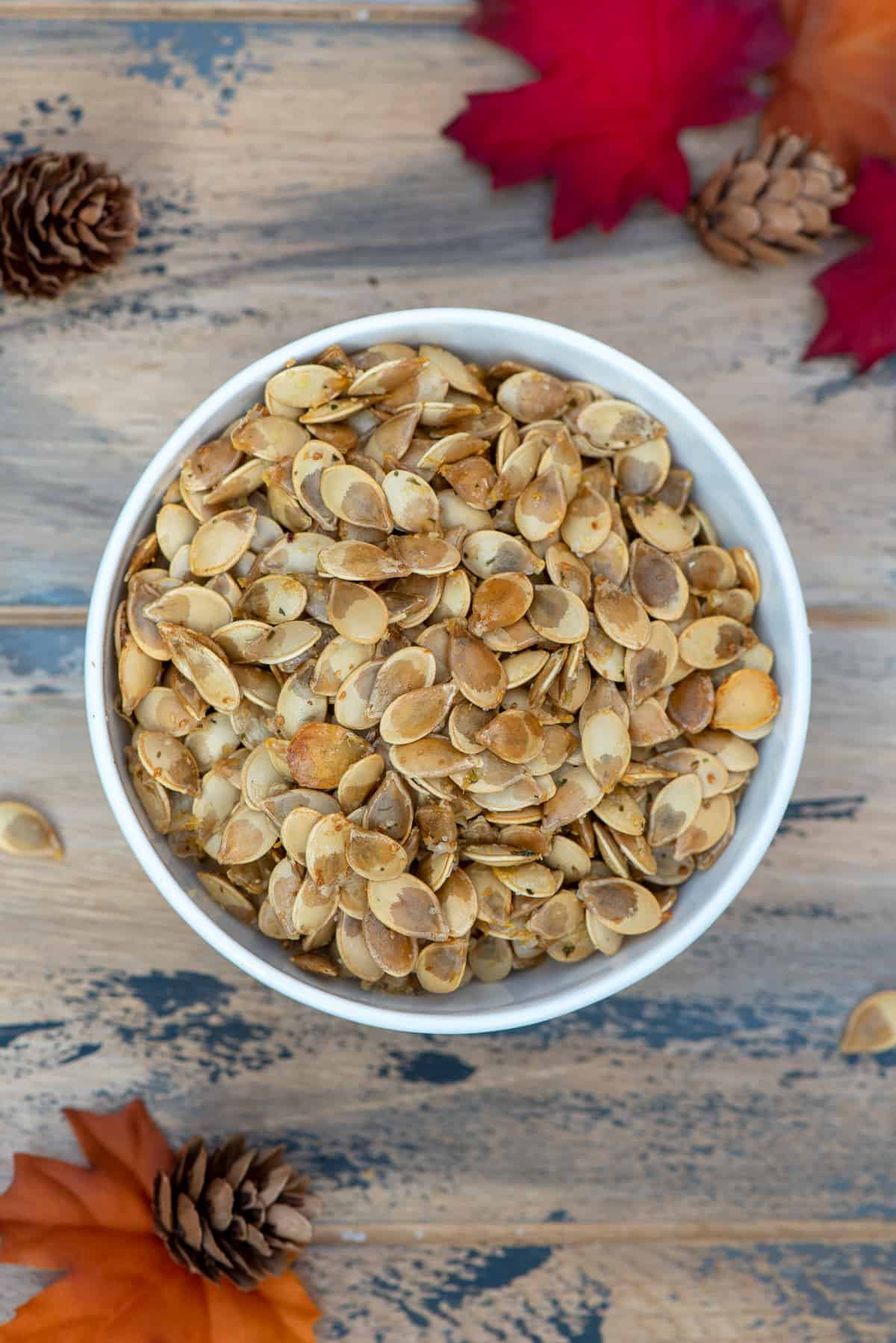 A top down shot of a bowl of roasted acorn squash seeds on a brown ball with fall leaves and pine cones.