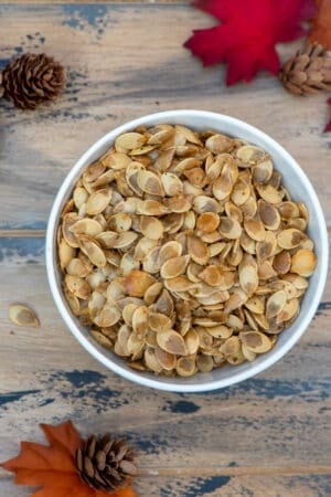 A top down shot of a bowl of roasted acorn squash seeds on a brown ball with fall leaves and pine cones.