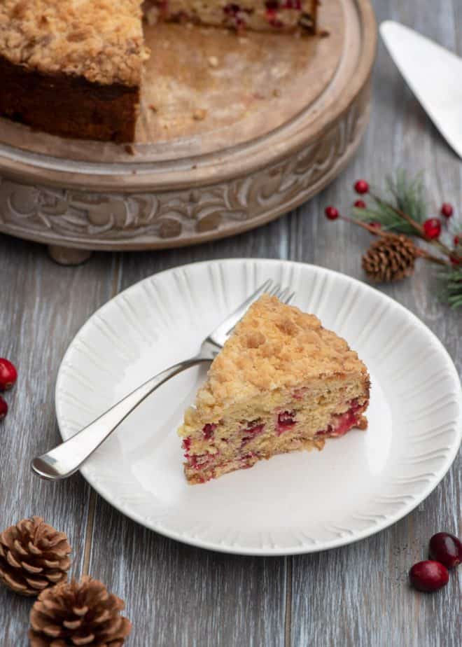 A slice of Cream Cheese Cranberry Coffee Cake on a white plate with a fork with pine cones scattered around.