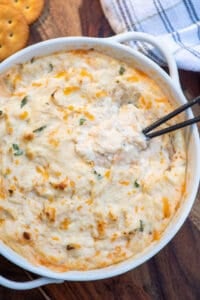 A top down shot of a small spoon resting in a white bowl filled with cheesy crab dip.