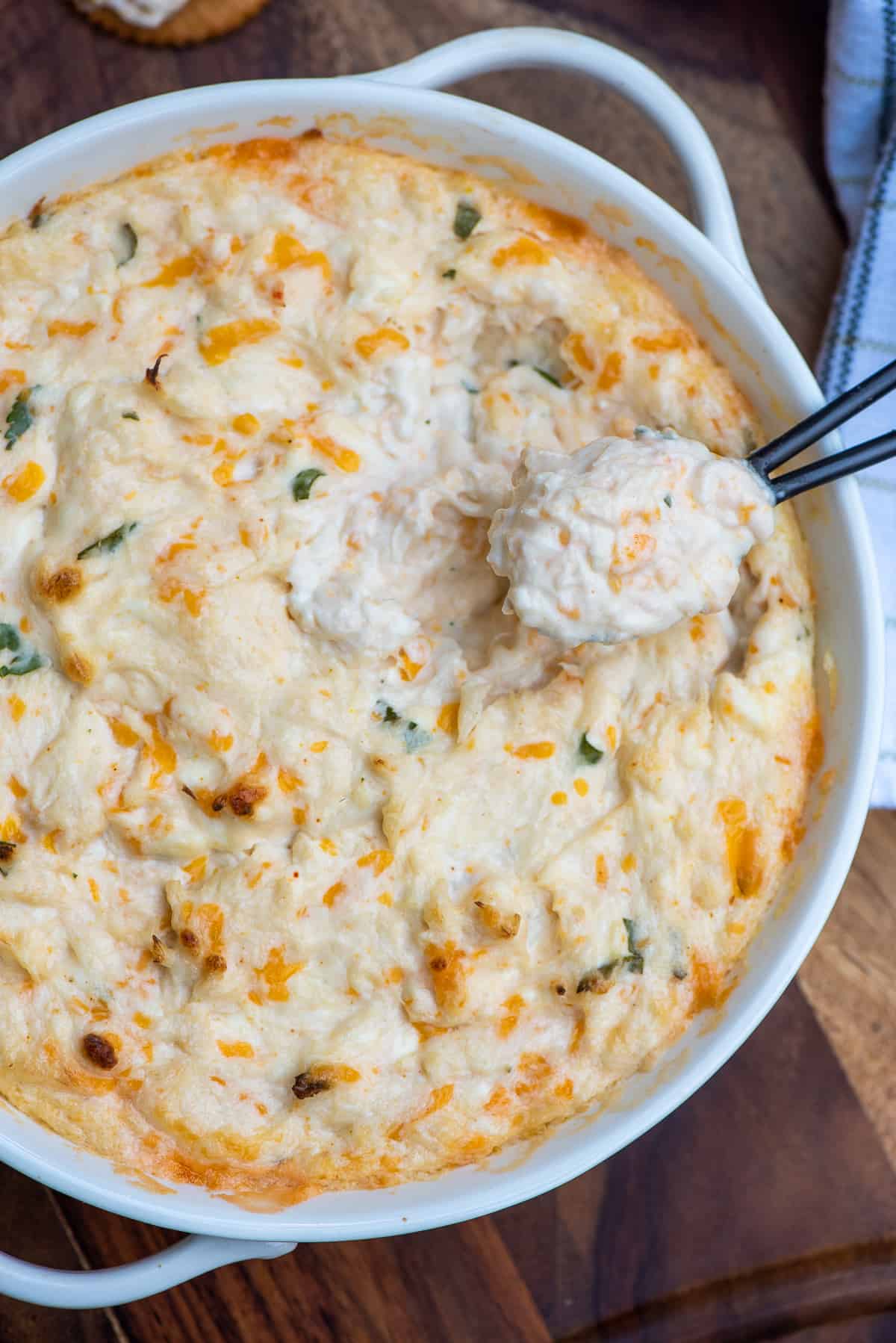 A small spoon scoops cheesy hot crab dip from a white bowl.