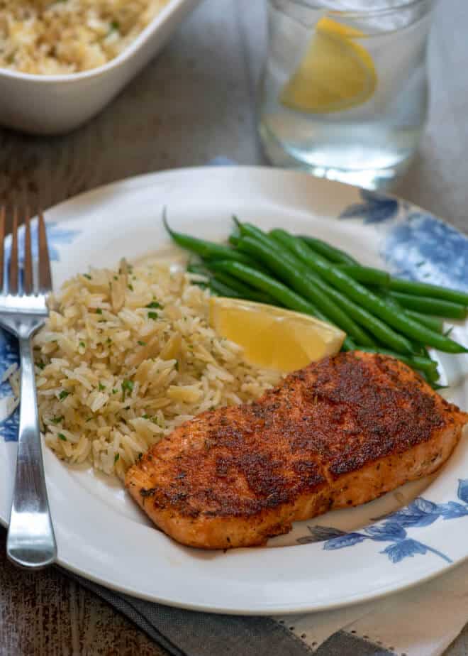 A piece of Blackened Salmon on a white plate with rice pilaf, green beans, and a slice of lemon..