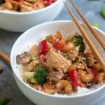 A bowl of cooked cubed chicken with red peppers, broccoli, and cashews with chopsticks lying over the top.