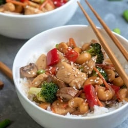 A bowl of cooked cubed chicken with red peppers, broccoli, and cashews with chopsticks lying over the top.