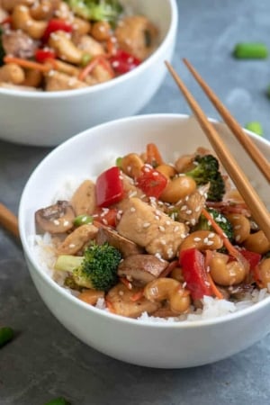 A bowl of cooked cubed chicken with red peppers, broccoli, and cashews with chopsticks lying over the top.