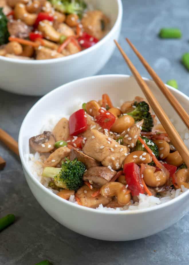 A bowl of cooked cubed chicken with red peppers, broccoli, and cashews with chopsticks lying over the top.