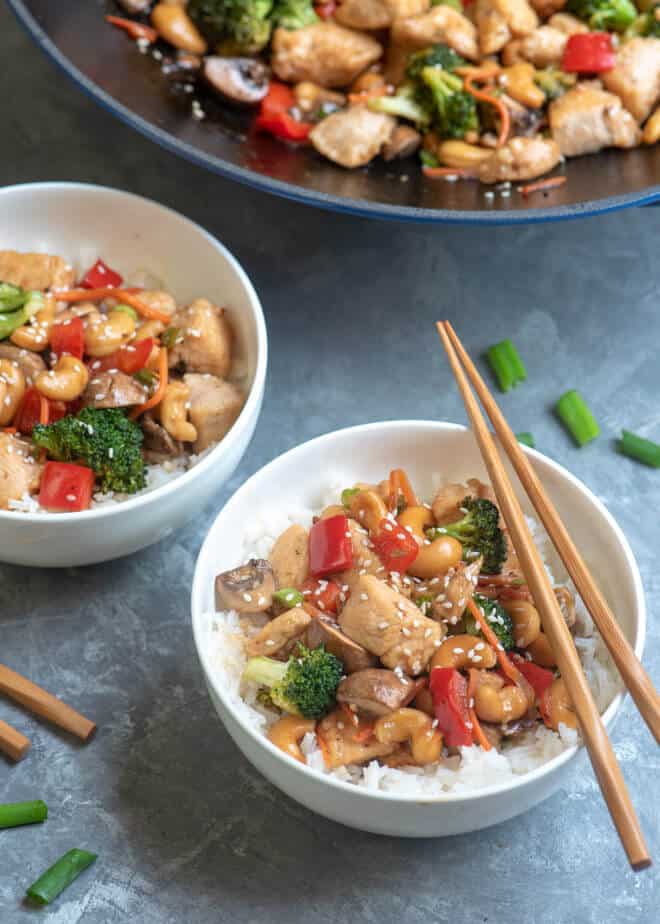 Two white serving bowls filled with Cashew Chicken with the wok in the backgroun.