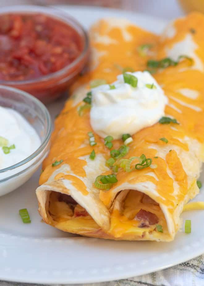 A close up image of the Ham and Cheese Overnight Breakfast Enchiladas on a white plate topped with sour cream.