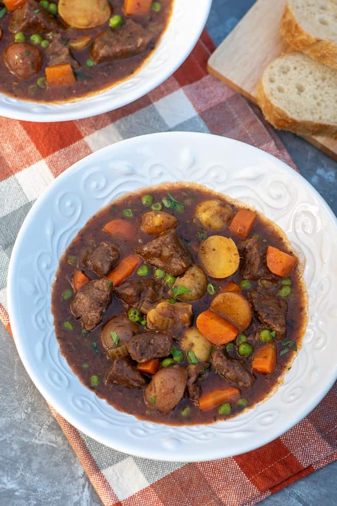 Beef Stew in a white serving bowl with sliced French bread in the background.