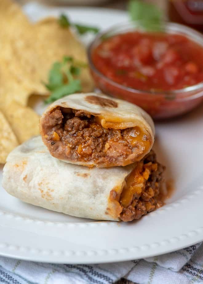 A close up of a beef and bean burrito cut in half on a white place with tortilla chips and a small bowl of salsa.