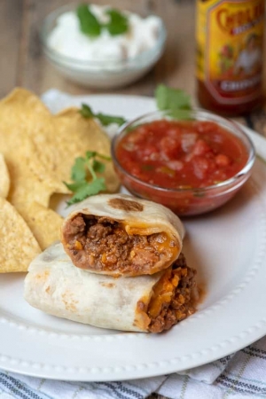 A burrito cut in half and stacked on a white plate with a small bowl of salsa and tortilla chips.