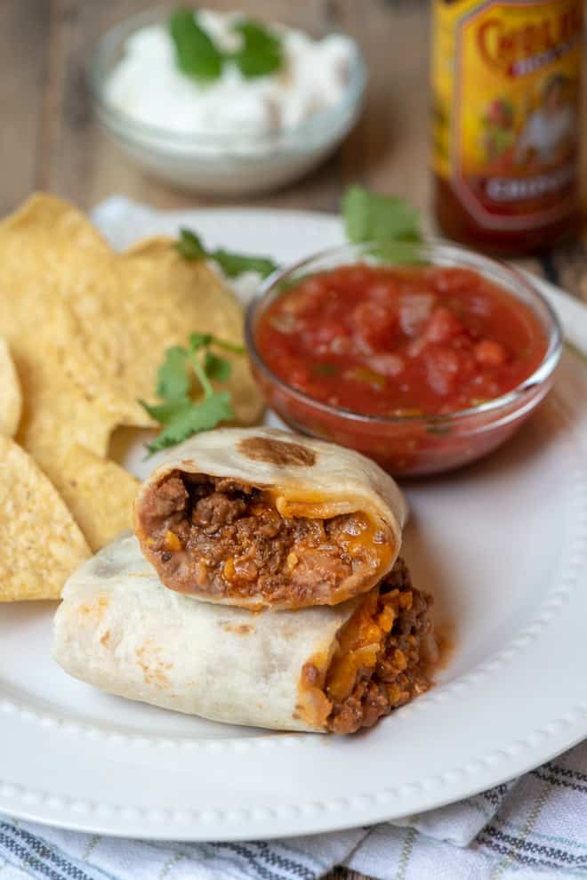 A burrito cut in half and stacked on a white plate with a small bowl of salsa and tortilla chips.