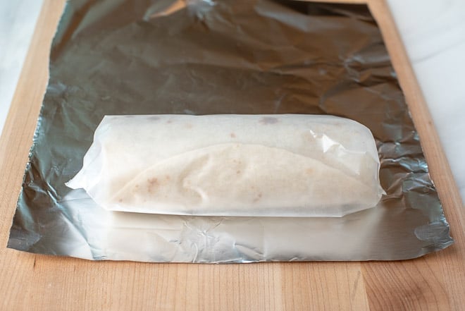 A burrito wrapped in wax paper on a sheet of foil.