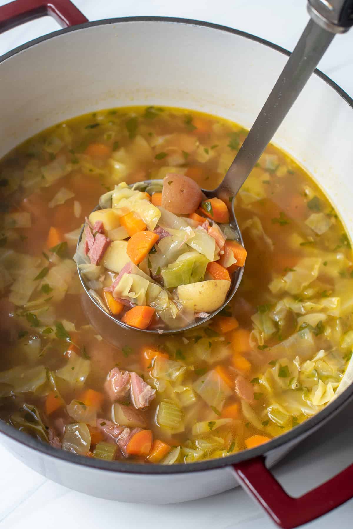 A ladle scooping corned beef and cabbage soup from a Dutch Oven.