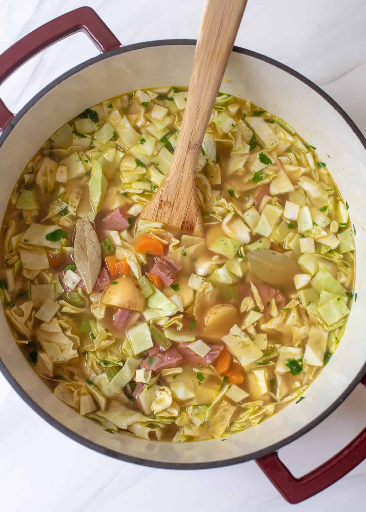 Corned beef and cabbage soup ingredients in a Dutch oven with a wooden spoon.