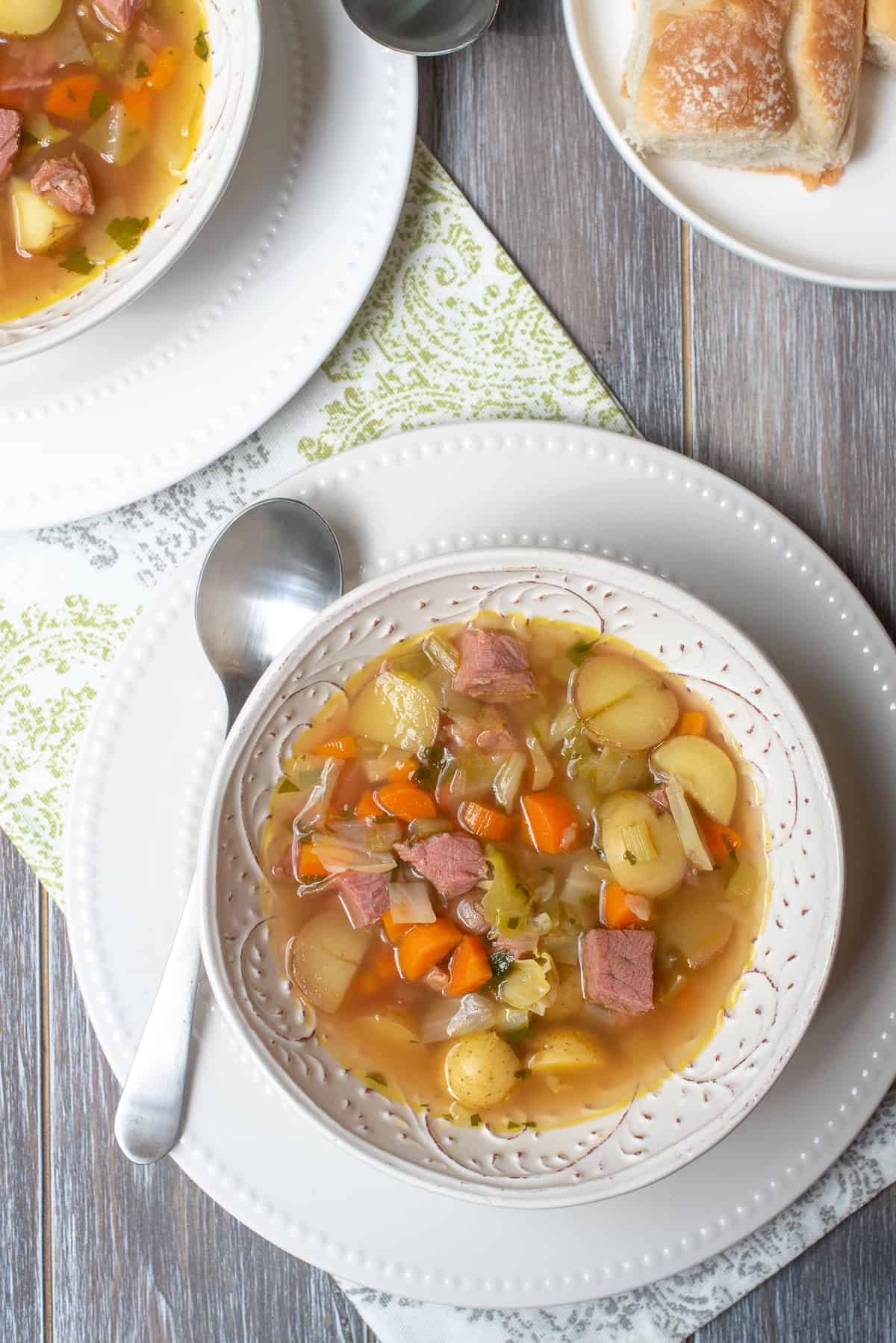 A bowl of soup with cabbage and corned beef on top of a white plate with a spoon.