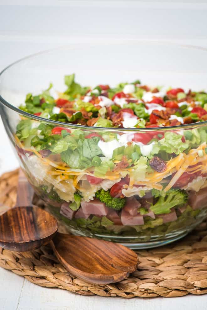 A glass bowl filled layers of bow tie pasta, greens, ham, and vegetables.