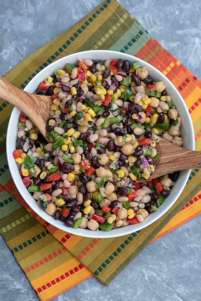 Mexican Three Bean Salad in a white serving bowl on a colorful cloth with wooden spoons in it.