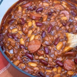 A pot of beans with kielbasa being stirred with a wooden spoon.
