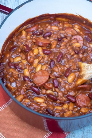 A pot of beans with kielbasa being stirred with a wooden spoon.