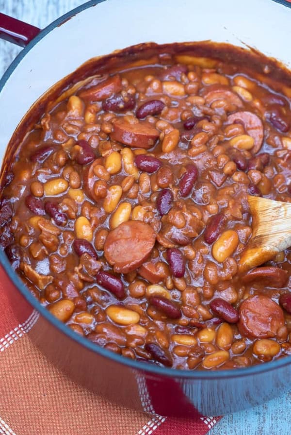 A pot of beans with kielbasa being stirred with a wooden spoon.