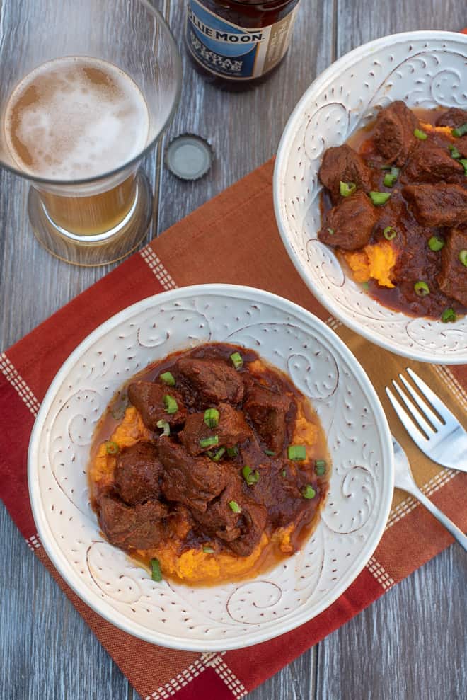 An overhead shot of two bowls of Smoky Chili Braised Beef with Sweet Potatoes on a patterned orange cloth with a beer.