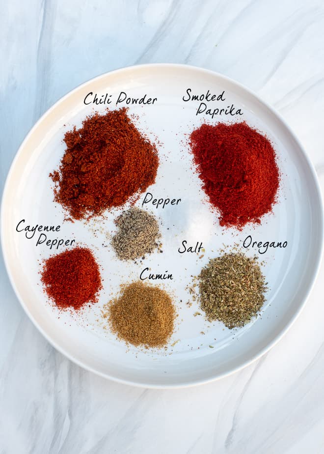 The spices that make up the Smoky Chili Seasoning Mix for the Carne Guisada on a white plate with text overlay - Chili Powder, Smoked Paprika, Oregano, Cumin, Cayenne Pepper, Salt, Pepper