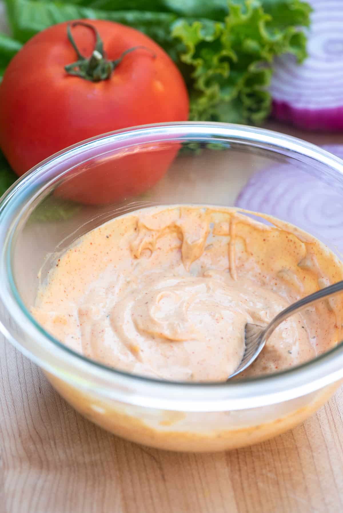 A bowl filled with burger sauce in front of a large red tomato and green lettuce leaves.