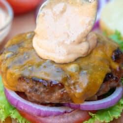 A spoon placing burger sauce on top of a cheeseburger.