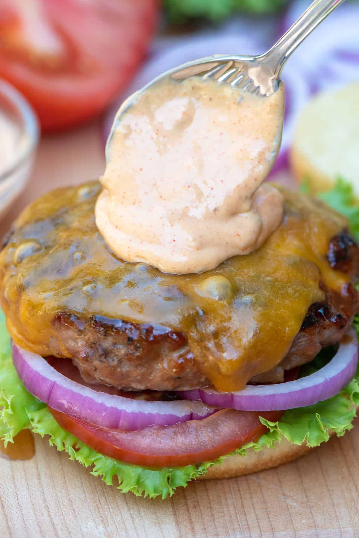 A spoon placing burger sauce on top of a cheeseburger.