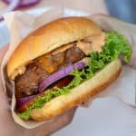 Hands holding a turkey burger with red onion, lettuce, and sauce wrapped in parchment paper.