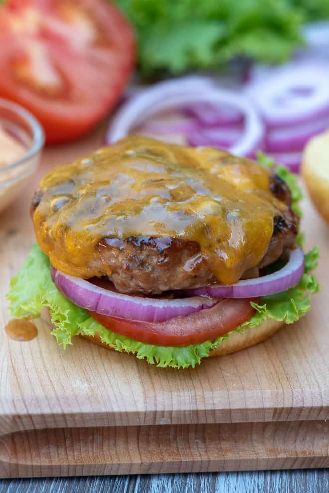 A cooked turkey burger with melted cheddar cheese rests on top of the bottom half of a bun with lettuce, sliced tomato, and red onion.