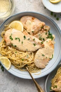 A top down shot of chicken scallopini on a plate with parmesan noodles and lemon wedges.