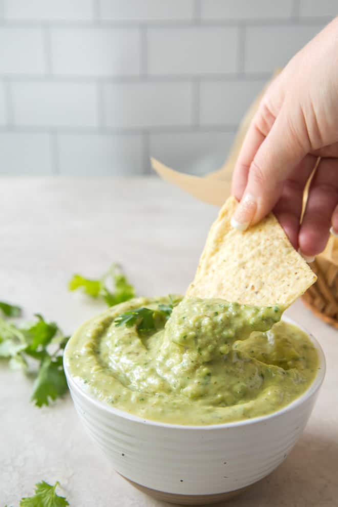 A hand dipping a chip into a white bowl full of creamy avocado salsa.
