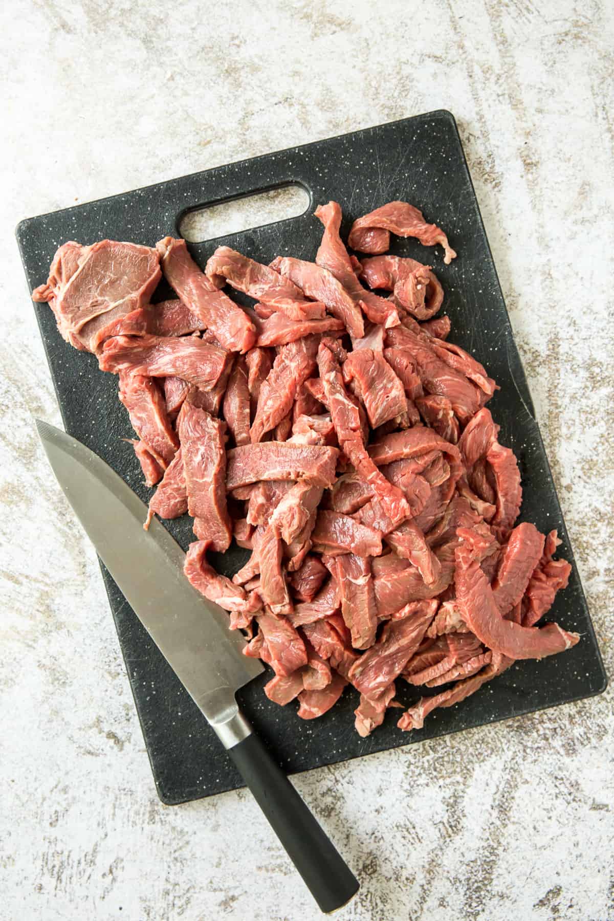 Thin strips of sirloin steak on a cutting board with a knife.