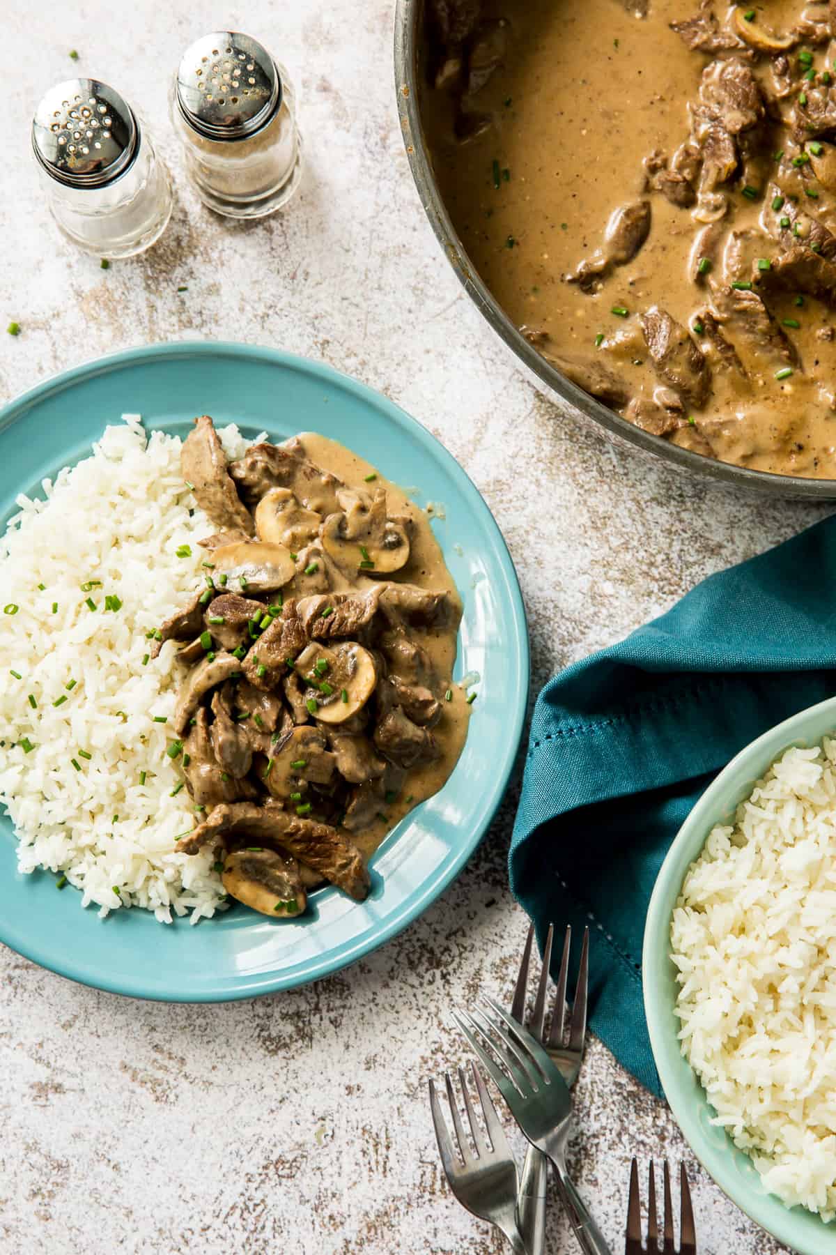 Beef stroganoff with mushrooms and wh white rice on a blue plate next to a skillet full of stroganoff.