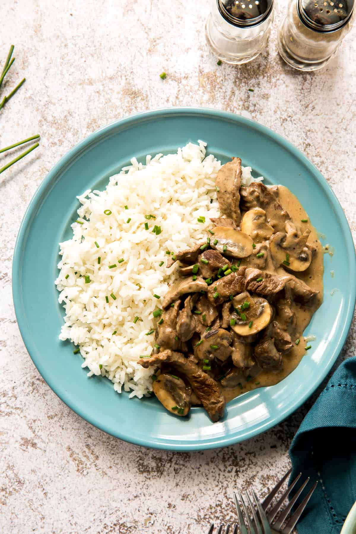 Beef stroganoff with mushrooms and wh white rice on a blue plate next to salt and pepper shakers.