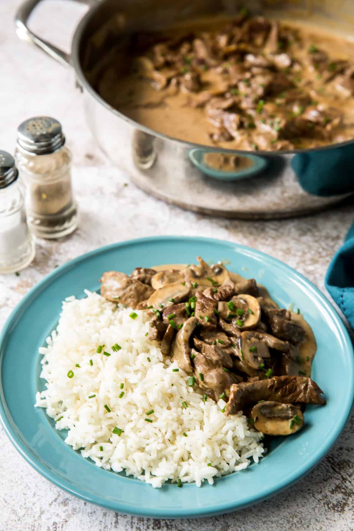 Beef stroganoff with mushrooms and wh white rice on a blue plate next to a skillet and salt and pepper shakers.