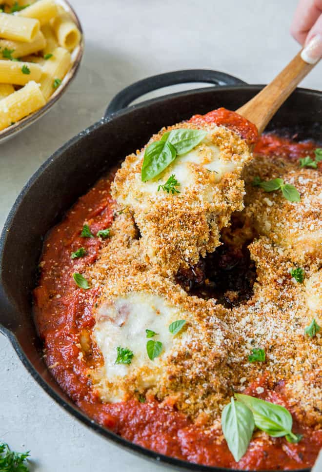 A wooden spoon scooping up a serving of Chicken Parmesan out of a cast iron skillet.