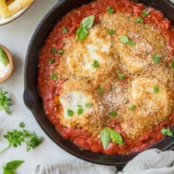 Chicken parmesan topped with breadcrumbs in a cast iron skillet next to a bowl of rigatoni pasta.