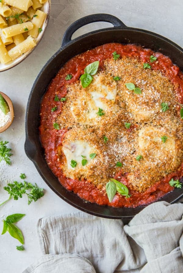 Chicken parmesan topped with breadcrumbs in a cast iron skillet next to a bowl of rigatoni pasta.