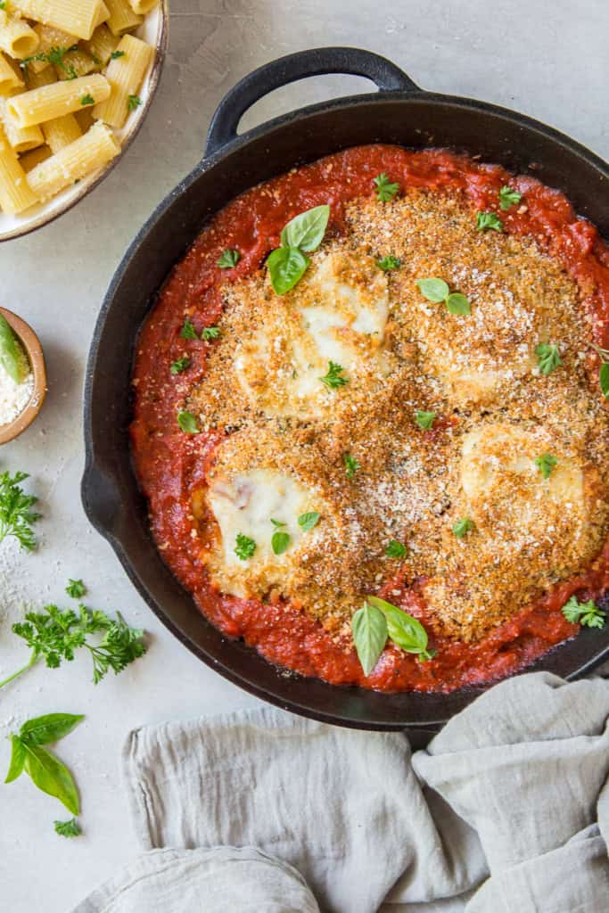 Chicken parmesan topped with breadcrumbs in a cast iron skillet next to a bowl of rigatoni pasta.