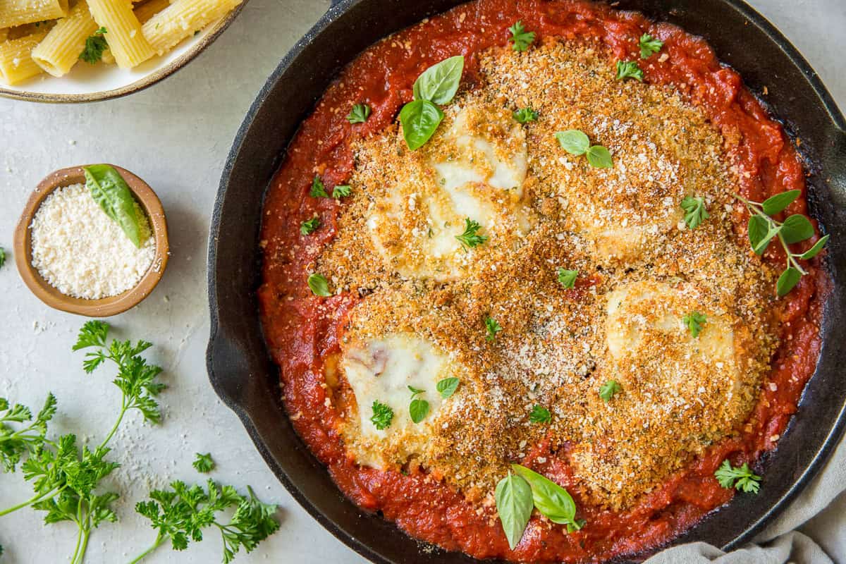 A small bowl of Parmesan next to a cast iron skillet filled with chicken parmesan in marinara sauce.