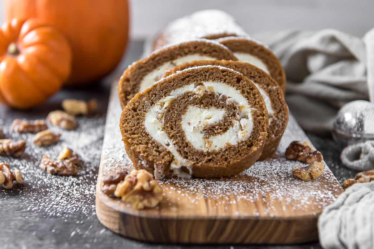 An eye level view of slices of pumpkin roll on a cutting board with small pumpkins and walnuts in the background.