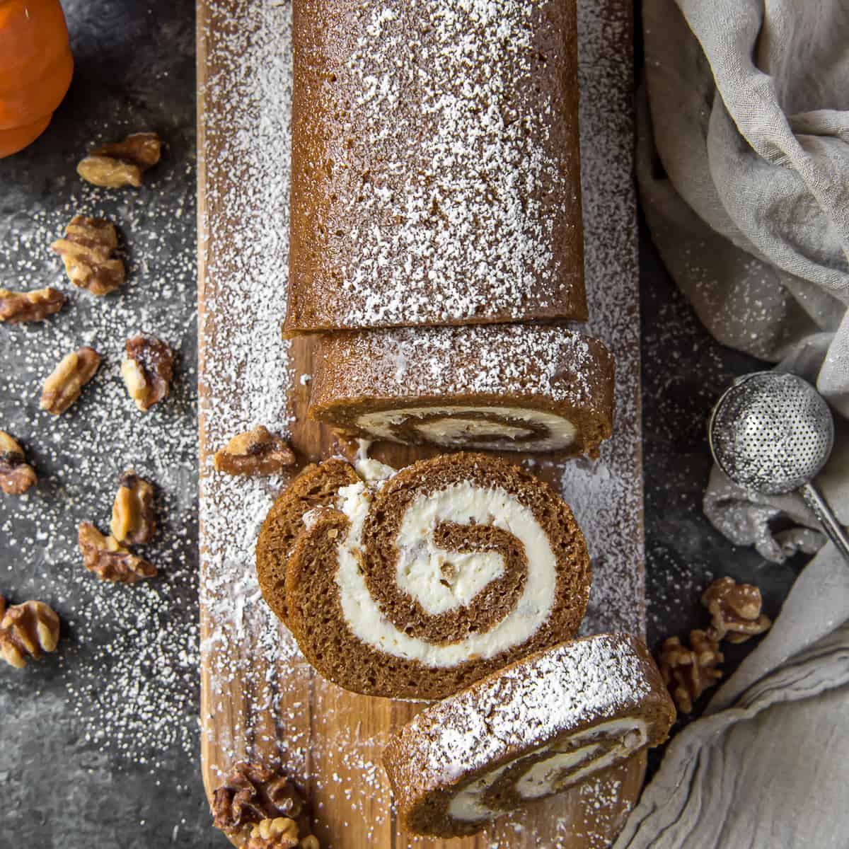 A top down shot of a partially sliced pumpkin roll coated with powdered sugar on a cutting board surrounded by walnuts.