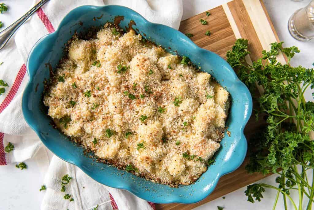 A blue scalloped baking dish filled with baked gnocchi covered in a breadcrumb topping, garnished with chopped parsley. The dish is placed on a white cloth with red stripes, next to a wooden cutting board with fresh parsley on it.