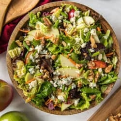 A top down shot of a cabbage salad with apples, blue cheese, and walnuts, a wooden salad bowl next to a red cloth and salad spoons.