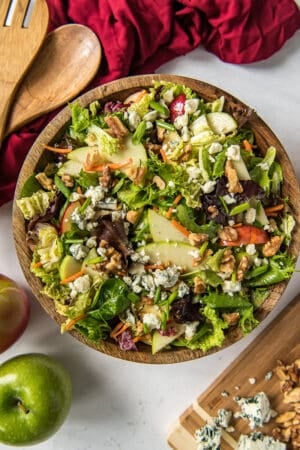 A top down shot of a cabbage salad with apples, blue cheese, and walnuts, a wooden salad bowl next to a red cloth and salad spoons.